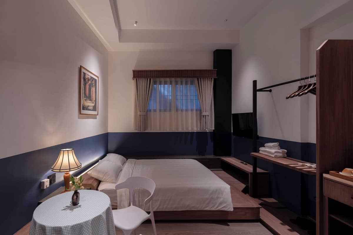 Une chambre d’hôtel comprenant un lit, une table de chevet avec une lampe, une chaise, une fenêtre avec des rideaux, un porte-manteau et une armoire.