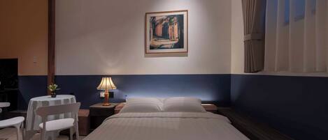 Habitación de hotel con cama, mesita de noche con lámpara, una pequeña mesa con silla y un cuadro en la pared.