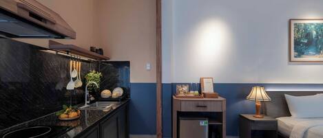 Una habitación de hotel compacta con kitchenette, una cama, una mesita de noche con lámpara y un cuadro en la pared.