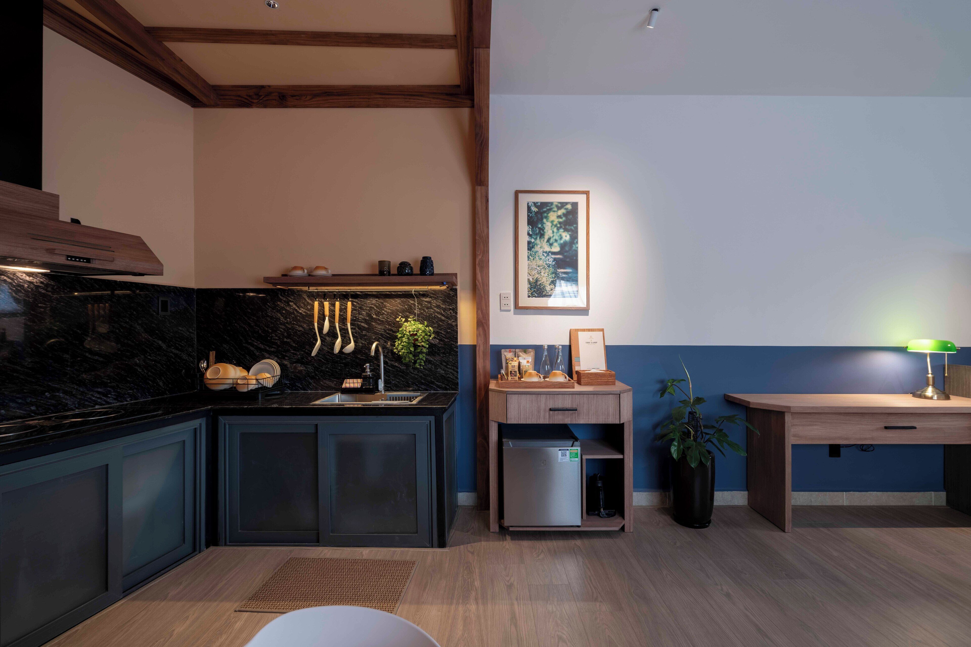 A modern kitchen with dark cabinetry, a marble backsplash, and a built-in sink. There is a small refrigerator and a wooden sideboard with a lamp.