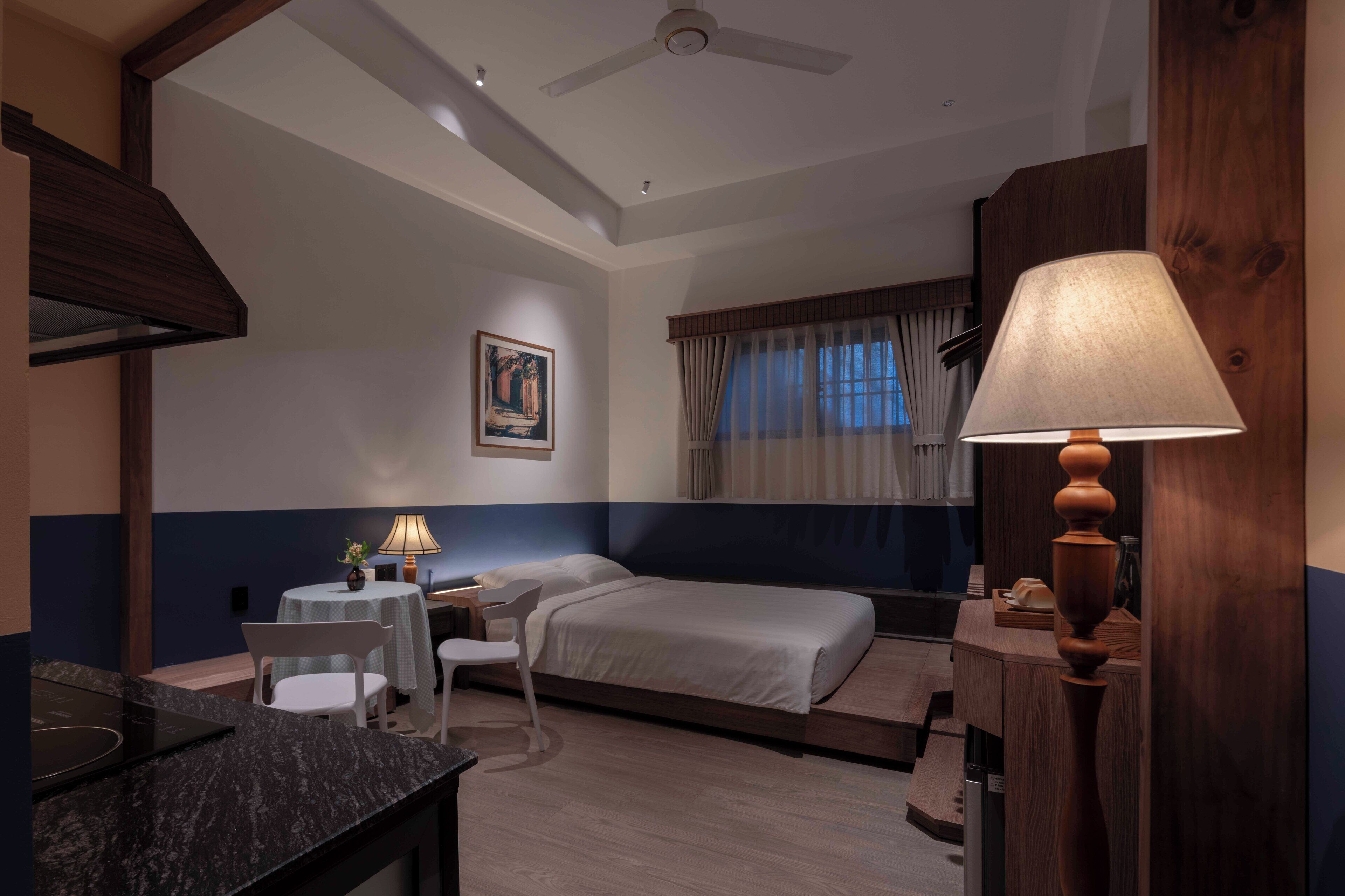 A hotel room with a bed, a desk with a chair, a small table with a lamp, and a painting on the wall.
