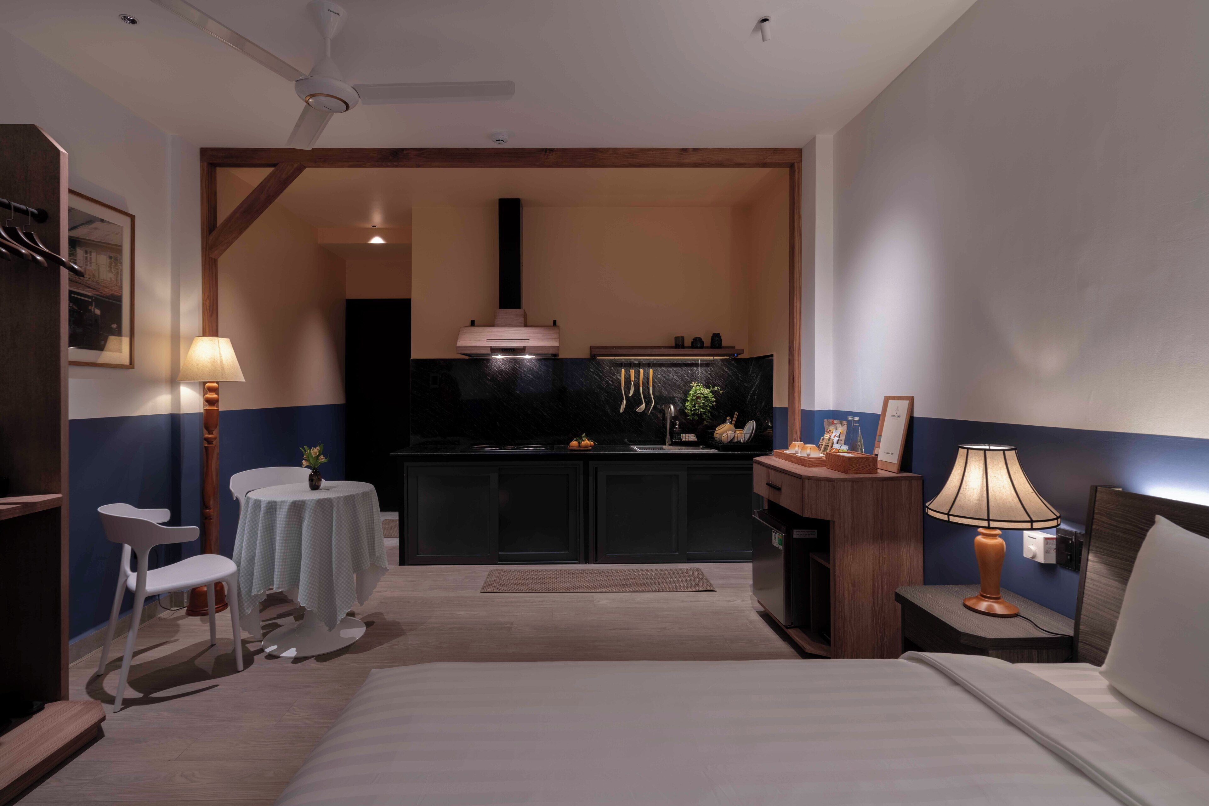 Une chambre d’hôtel comprenant un lit, un bureau, une chaise, une petite table, une lampe, une table de chevet, un téléviseur fixé au mur et un coin cuisine avec des placards et une cuisinière.