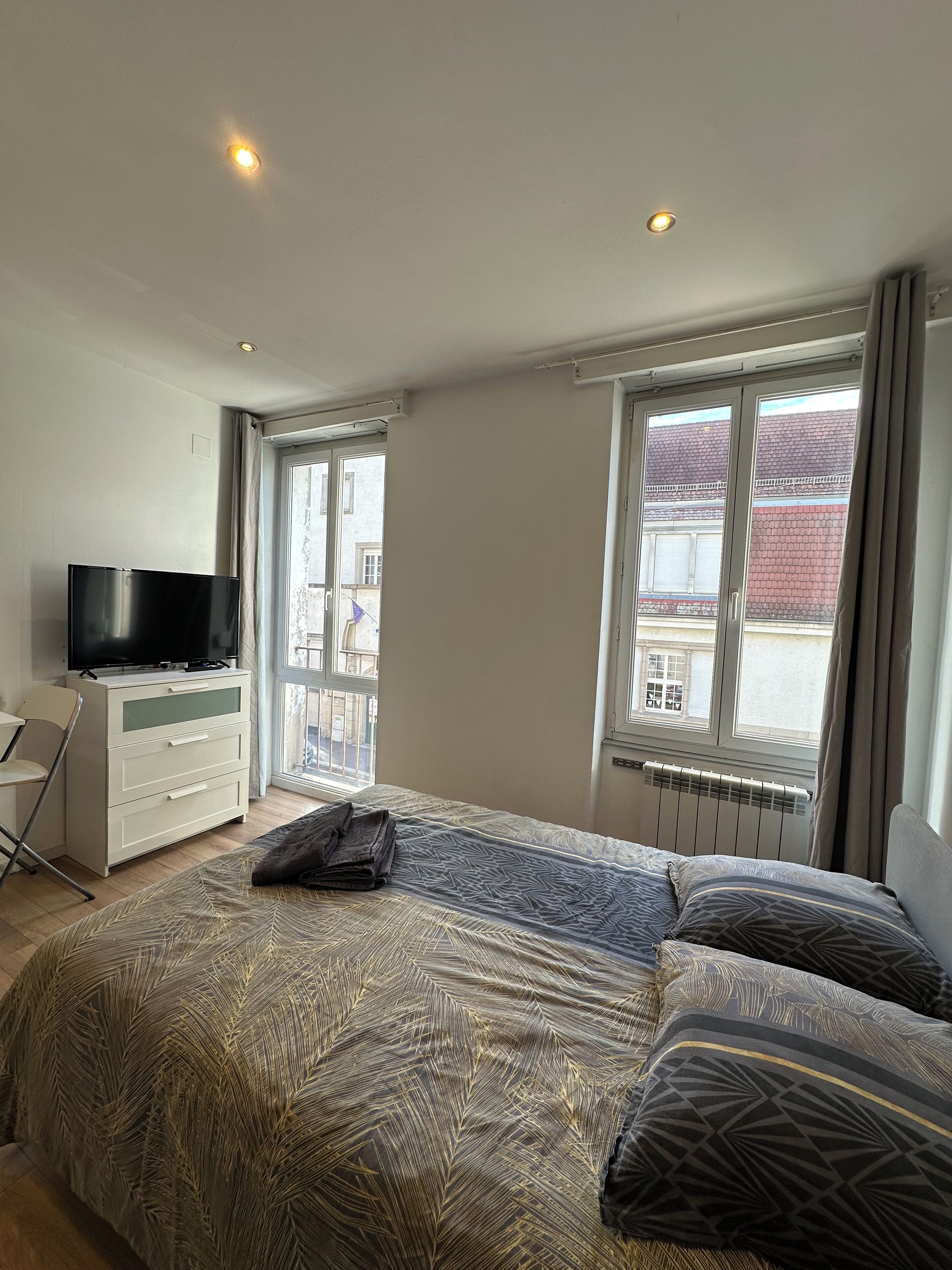 Une chambre à coucher comprenant un lit, une télévision, un bureau et une fenêtre donnant sur des immeubles.