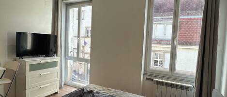 Une chambre à coucher comprenant un lit, une télévision, un bureau et une fenêtre donnant sur des immeubles.