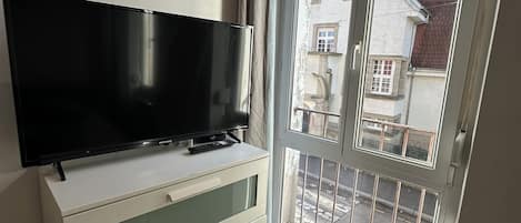 Una habitación con televisión de pantalla plana sobre un tocador blanco, puerta corrediza de vidrio a un balcón y vista de edificios.