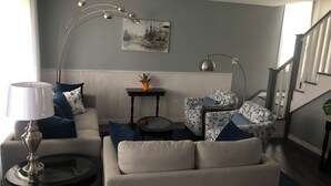 A living room with a sofa, armchair, and a coffee table. There is a painting on the wall and a staircase in the background. - Dragon Fly Lane, St. Ann's Rest & Relax or Explore in the very private setting . (Baddeck)