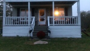 A white two-story house with a porch, a front door, and a small front yard. - Dragon Fly Lane, St. Ann's Rest & Relax or Explore in the very private setting . (Baddeck)