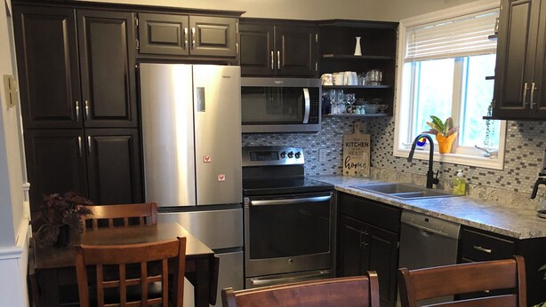 A modern kitchen with dark cabinetry, stainless steel appliances, and a dining area with wooden furniture. - Dragon Fly Lane, St. Ann's Rest & Relax or Explore in the very private setting . (Baddeck)
