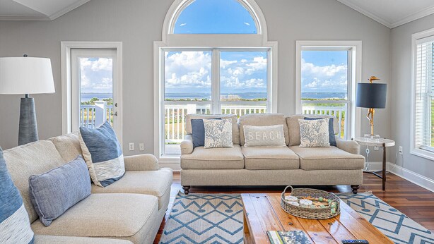 Amplia sala de estar con techo alto, ventanas grandes que ofrecen vista al mar, un sofá beige con cojines estampados, una mesita de noche con una lámpara y una mesa de centro de madera con una cesta.