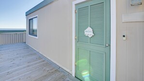Une terrasse en bois mène à une porte verte ornée d’une pancarte « Bienvenue », avec une petite fenêtre et un luminaire fixé au mur.