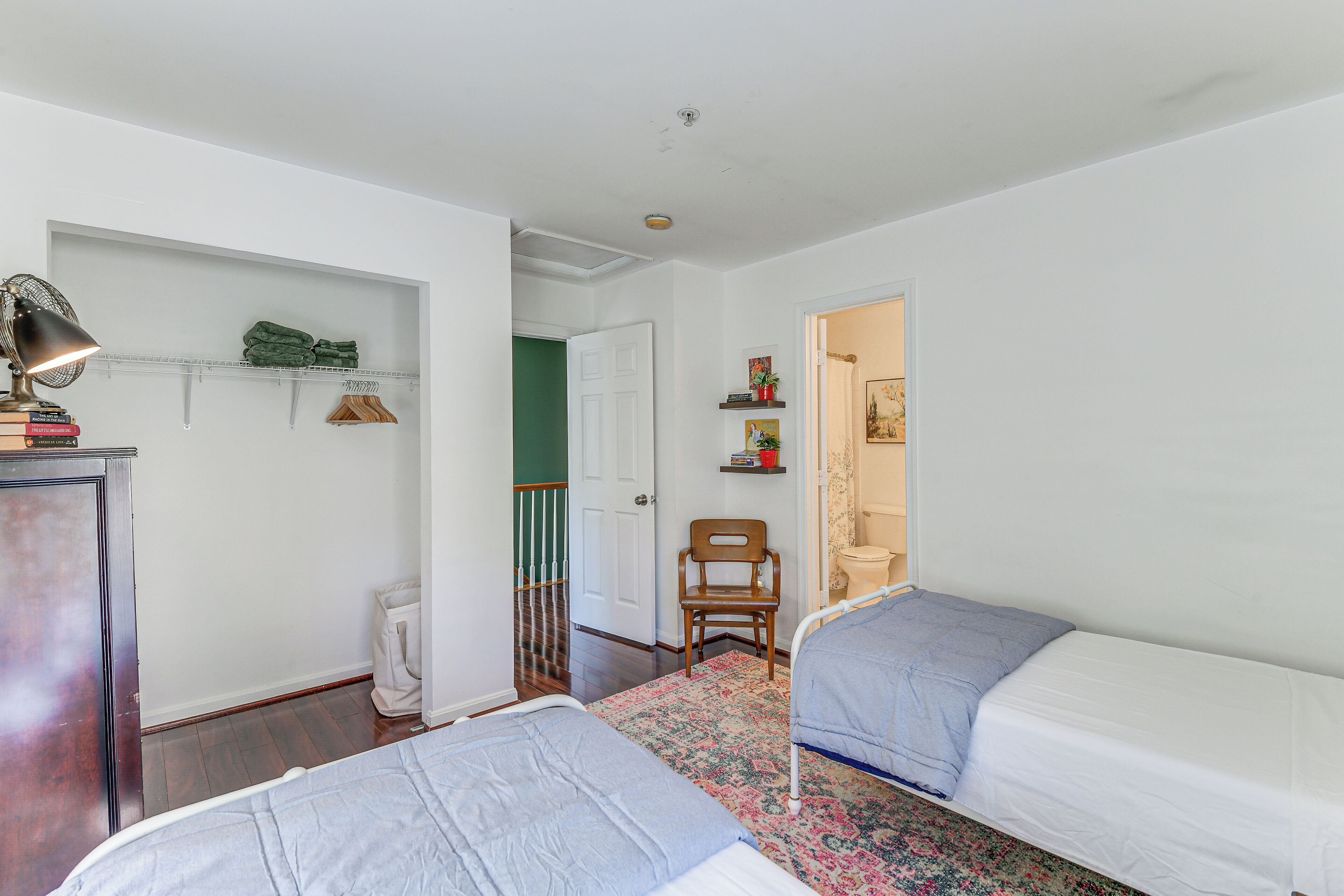 A bedroom with two beds, a desk with a lamp, a chair, a shelf with books and a plant, and a door leading to another room.