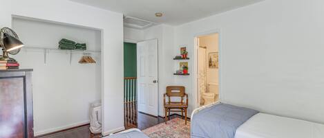 A bedroom with two beds, a desk with a lamp, a chair, a shelf with books and a plant, and a door leading to another room.