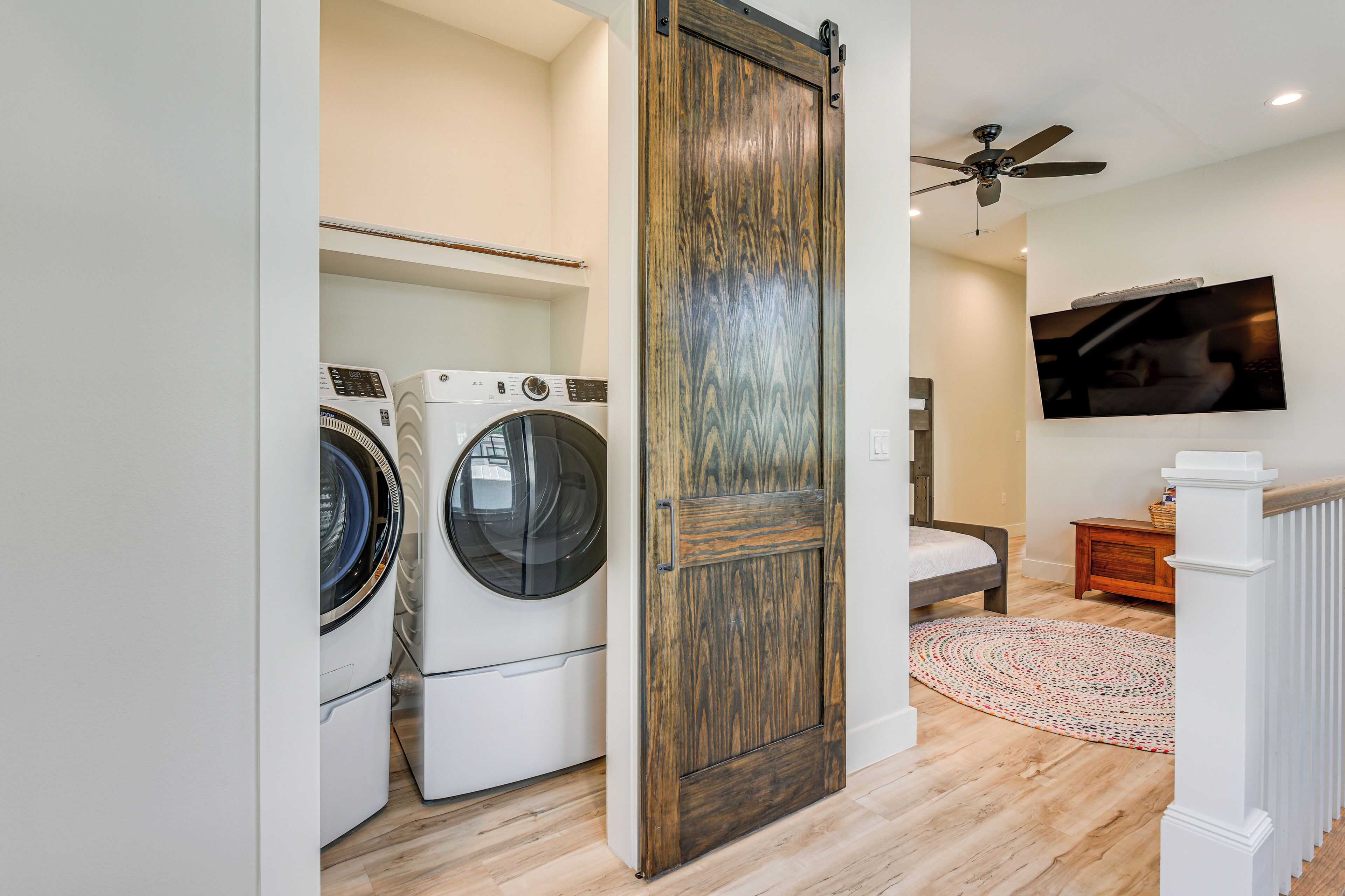 A laundry room with two front-loading washers and dryers, a wooden door, a ceiling fan, a television mounted on the wall, and a patterned rug on the floor.