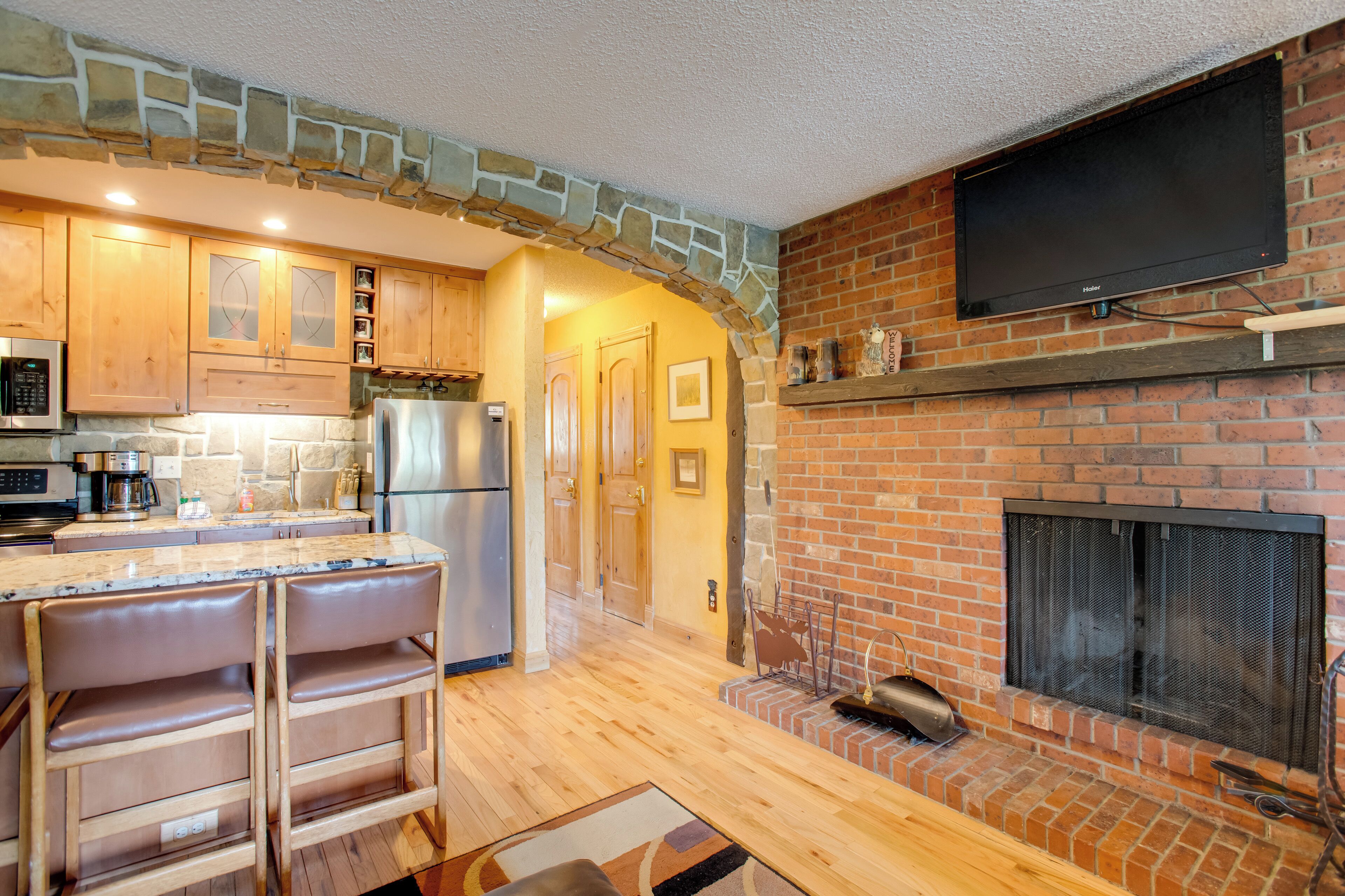A kitchen with a stone arch, wooden cabinets, a built-in stove, and a flat-screen TV mounted above the fireplace.