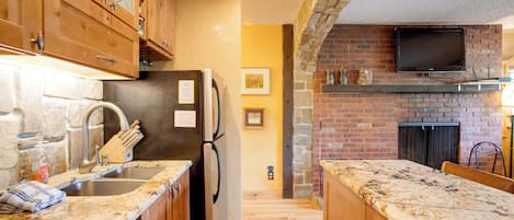 A kitchen with wooden cabinets, a granite countertop, a stainless steel sink, a black refrigerator, a brick wall, and a flat-screen TV mounted above the fireplace.