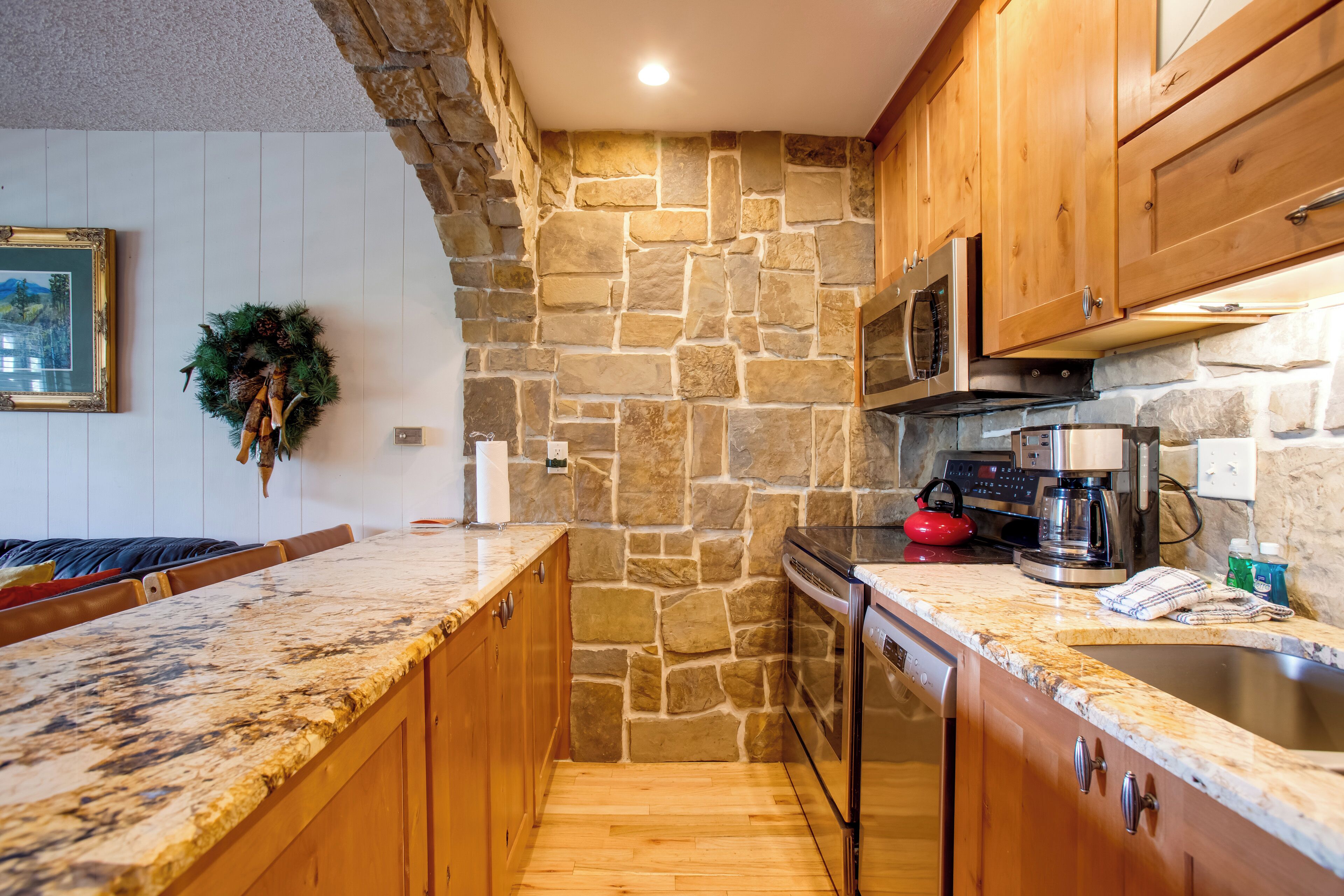 Una cocina con armarios de madera, pared de piedra, encimera de granito, microondas, cafetera y lavavajillas.