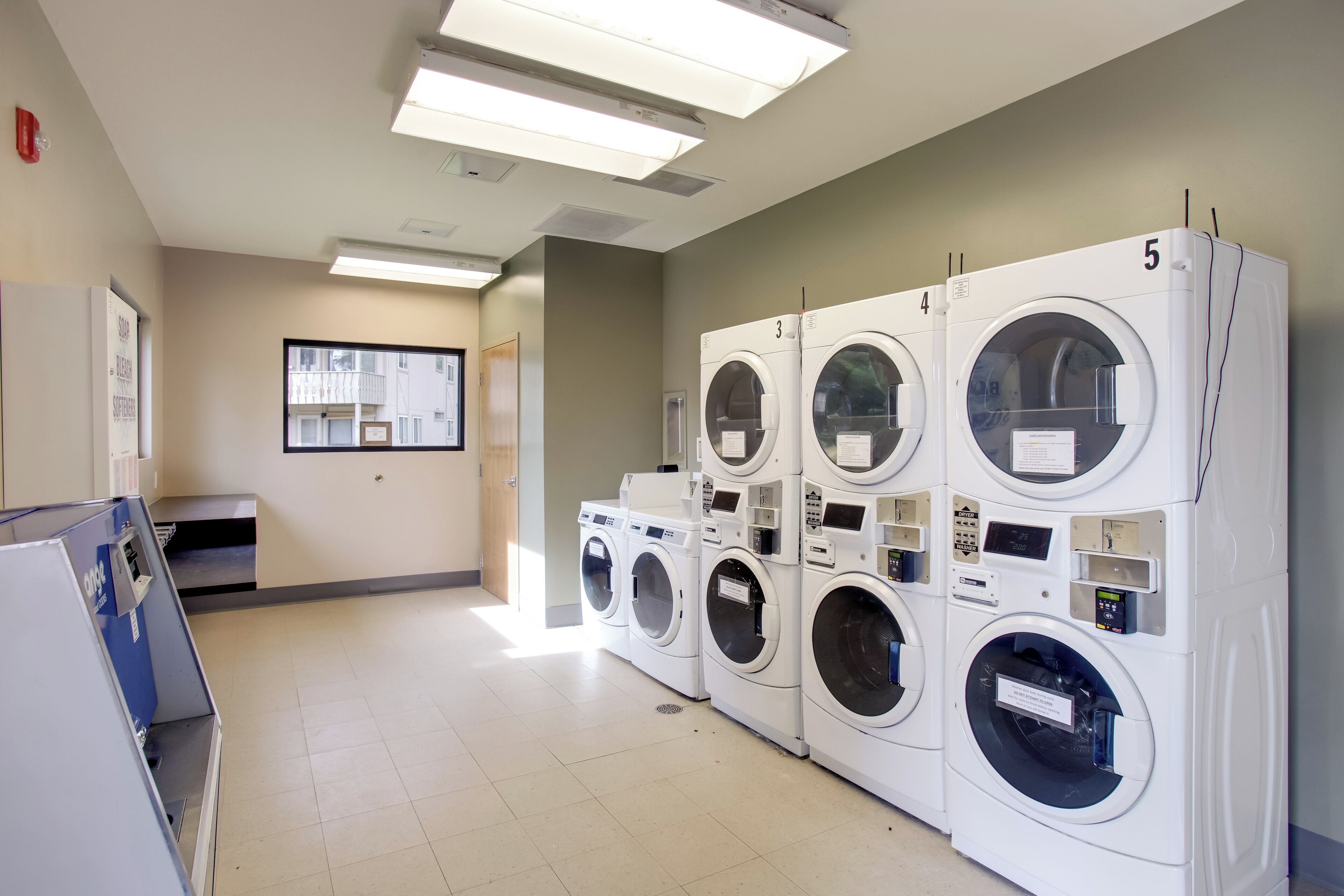 A laundry room with multiple washing machines, a control panel, and a window with a view of the outside.