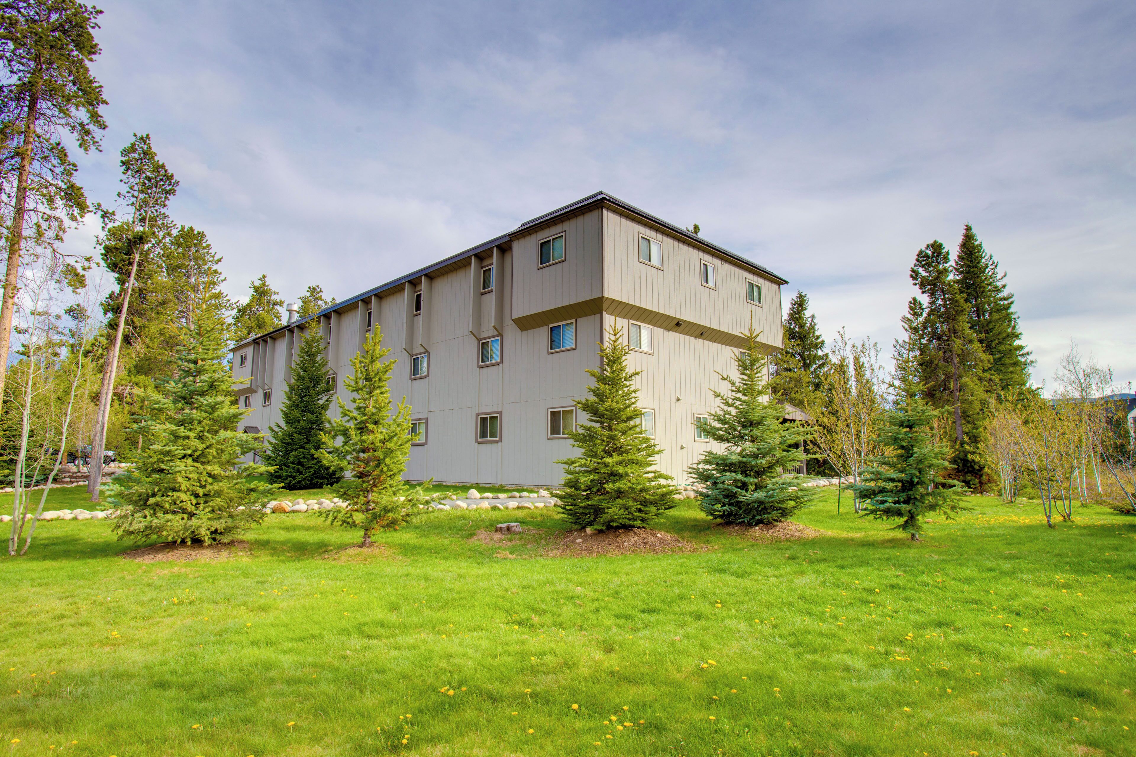 A multi-story building with a flat roof, surrounded by a grassy area with several evergreen trees.