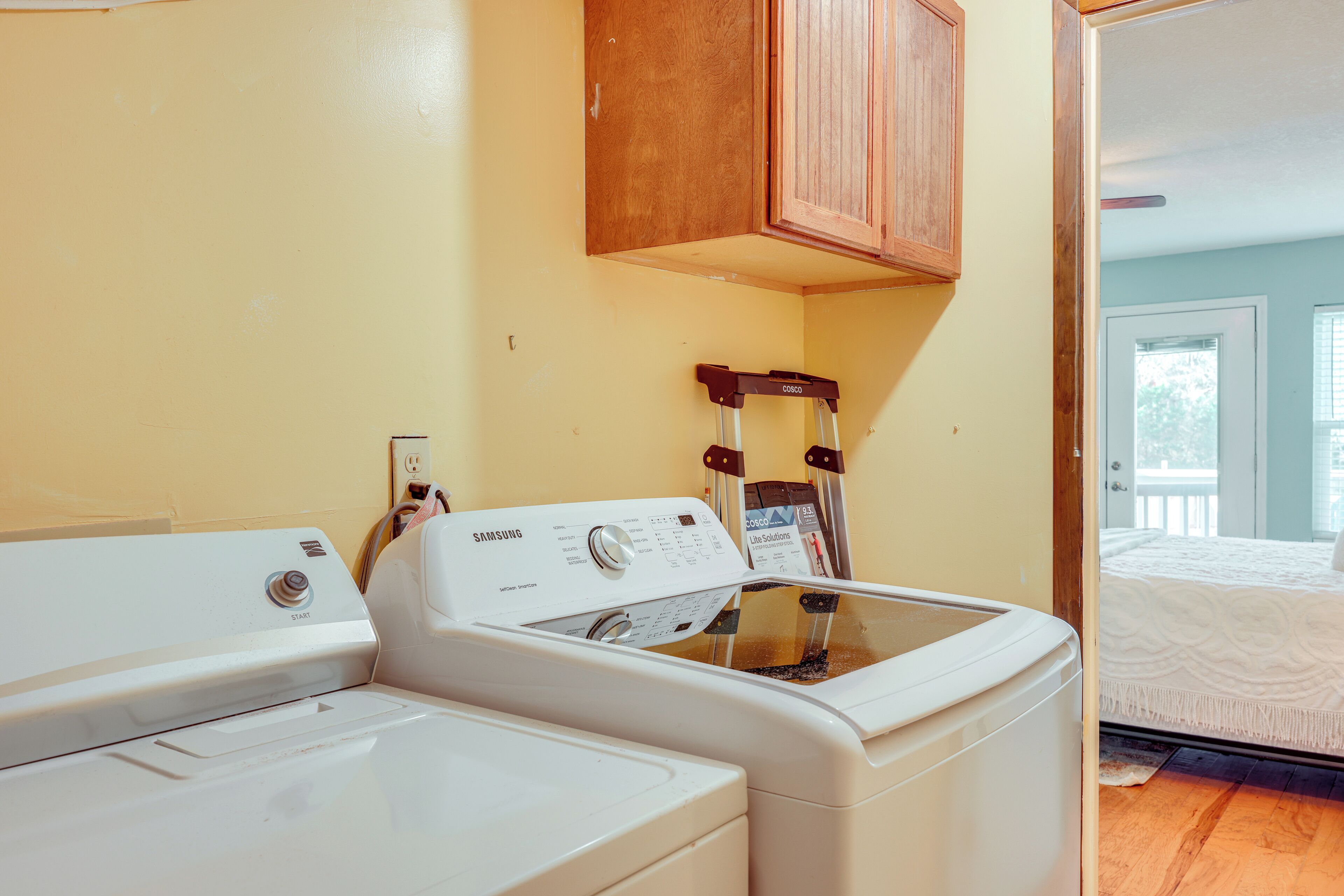 A laundry area with two white washing machines, a wooden cabinet above, and a door leading to a bedroom with a bed and a window.