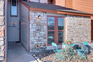 A stone exterior with a door labeled "Adventures Awaits," a small patio with chairs and a table, and a potted plant. - Hill Country Townhome: Close to Parks & Hiking (Wimberley)