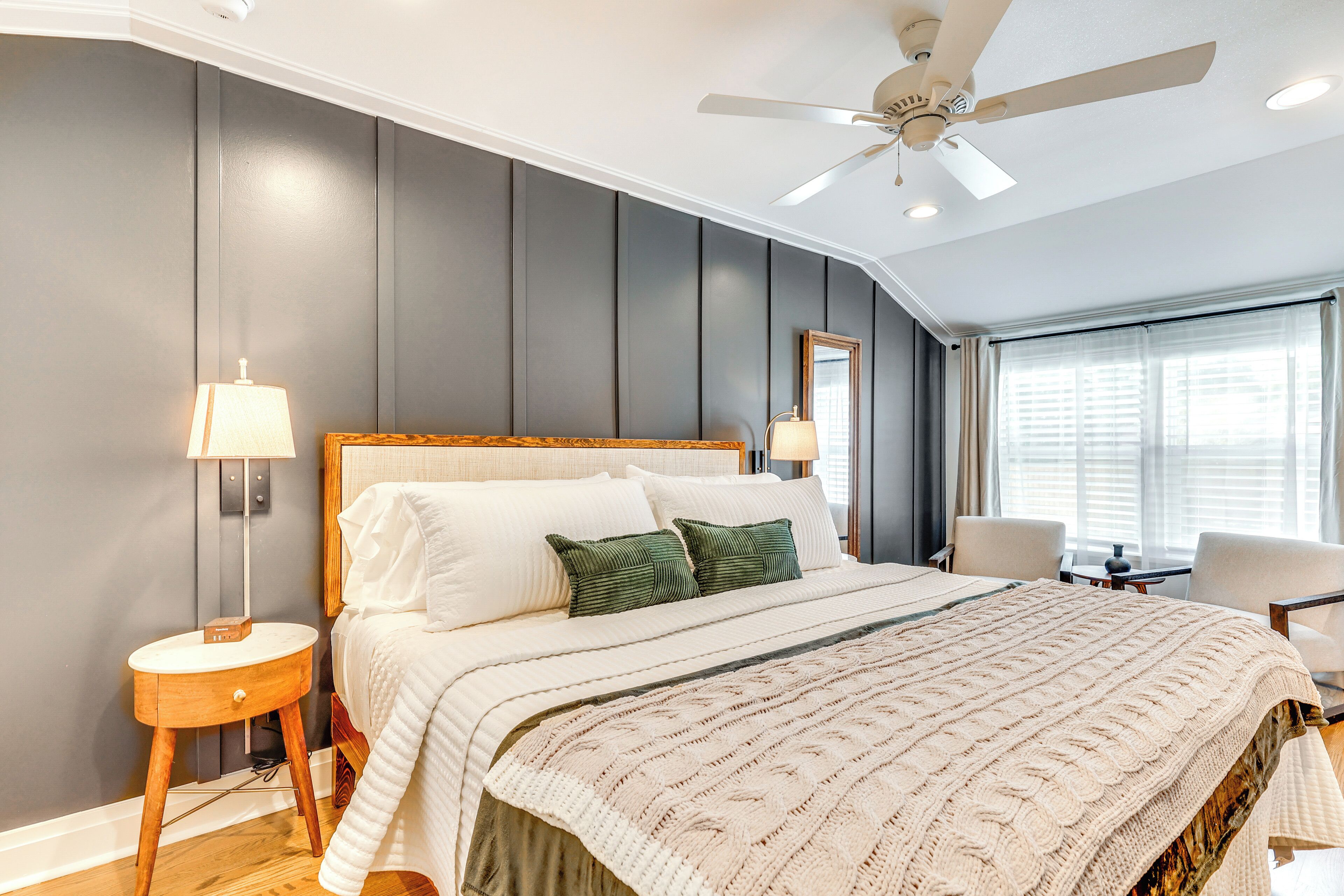 A bedroom with a large bed, two bedside lamps, a ceiling fan, and a view of the outside through a window.