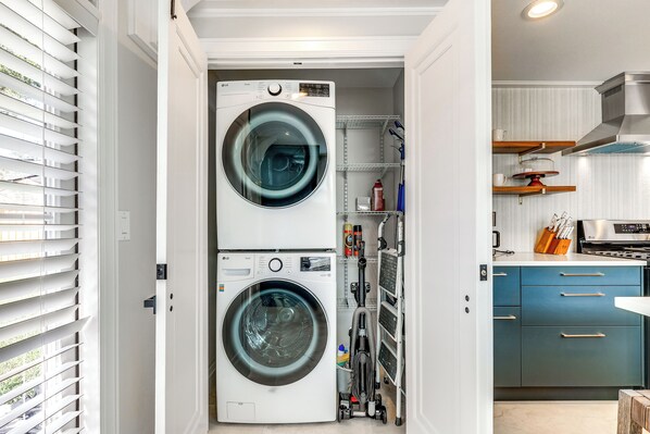 A laundry room with two stacked front-loading washing machines, a shelving unit with cleaning supplies, and a kitchen area with blue cabinets and a stove. - Fenced Yard: Lakeland Home Adjacent to Golf Course (Lakeland)
