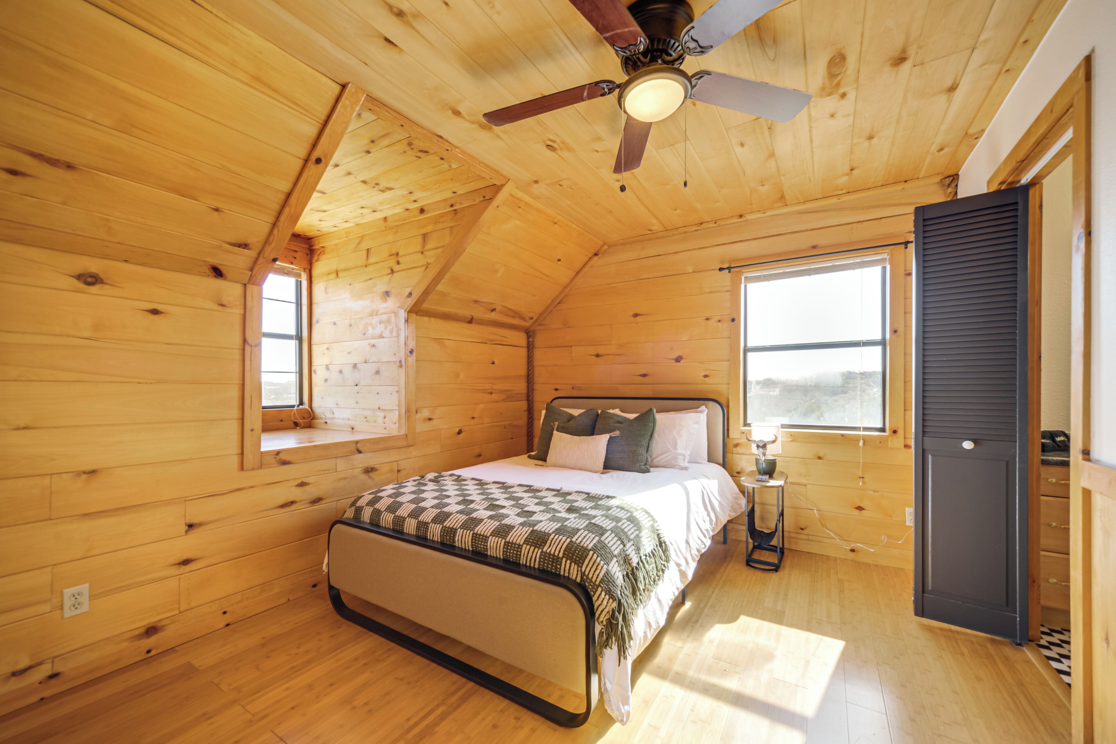 A wooden cabin bedroom with a bed, a ceiling fan, and two windows.