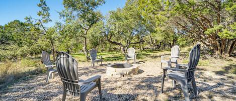 A patio area with Adirondack chairs arranged around a stone fire pit, surrounded by trees and gravel.