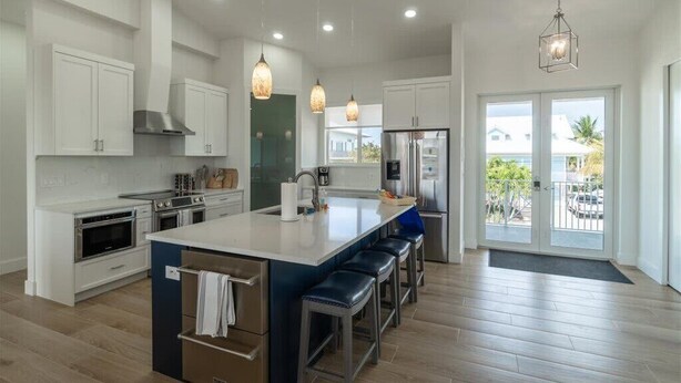 A modern kitchen with white cabinets, stainless steel appliances, a central island with bar stools, pendant lighting, and a view through the glass door. - Coco4Fun New Luxury Canal Front with Pool/Spa/Dock (Marathon)