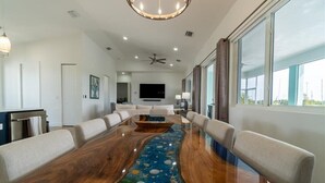 A modern living room with a large wooden dining table, white sofas, a flat-screen TV, and a large window. - Coco4Fun New Luxury Canal Front with Pool/Spa/Dock (Marathon)