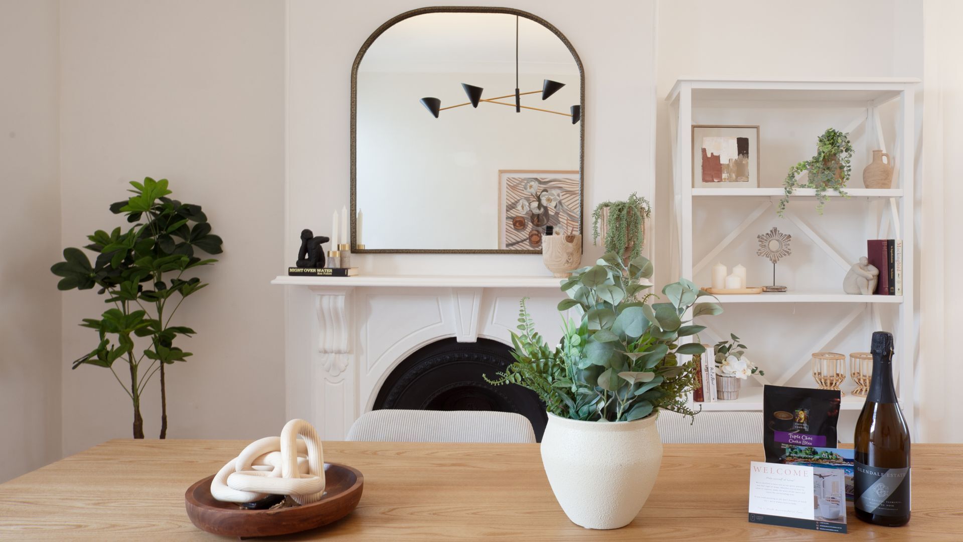 Une pièce avec une cheminée, une table en bois, une plante en pot, une bouteille de vin et une bibliothèque agrémentée d’objets décoratifs.