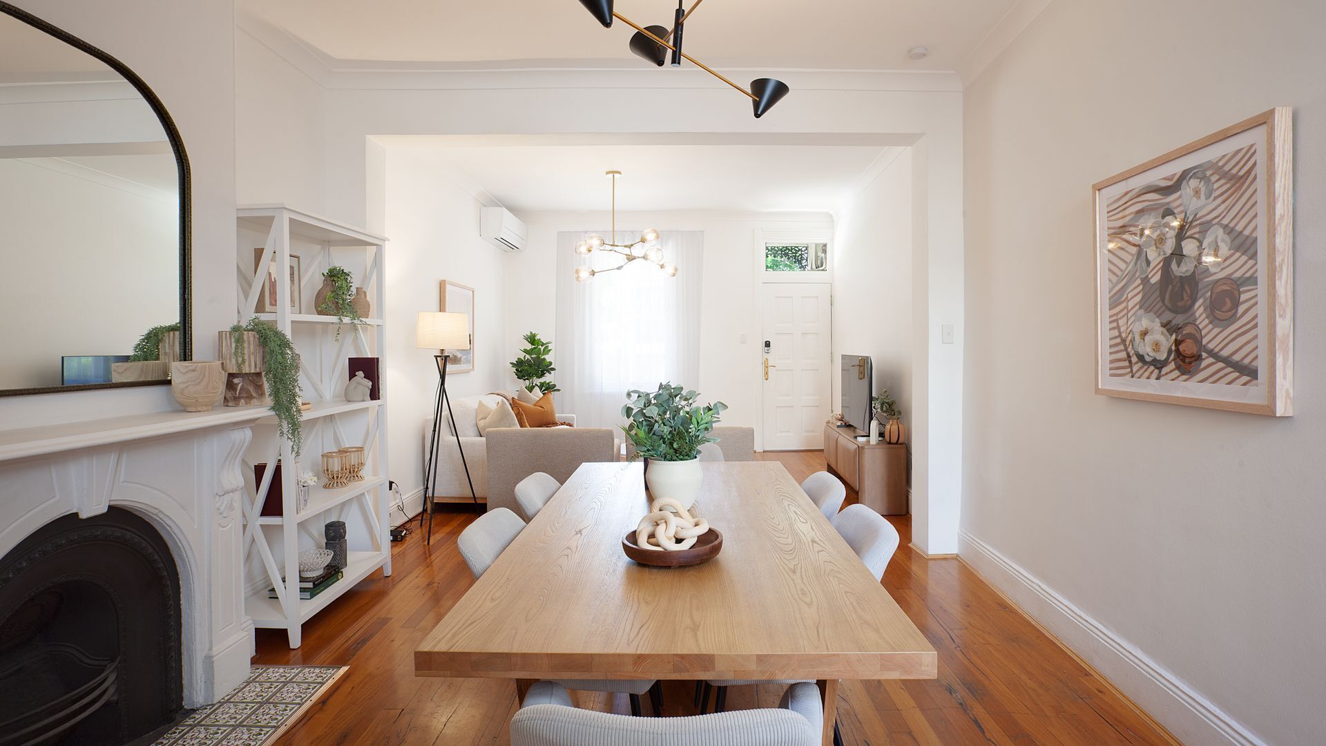 A spacious dining area with a wooden table, white chairs, and a fireplace. There is a bookshelf with plants and a floor lamp.