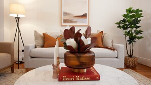 A living room with a round coffee table, a vase with flowers, a book titled 'Henri Matisse', a floor lamp, a sofa with cushions, and a potted plant.