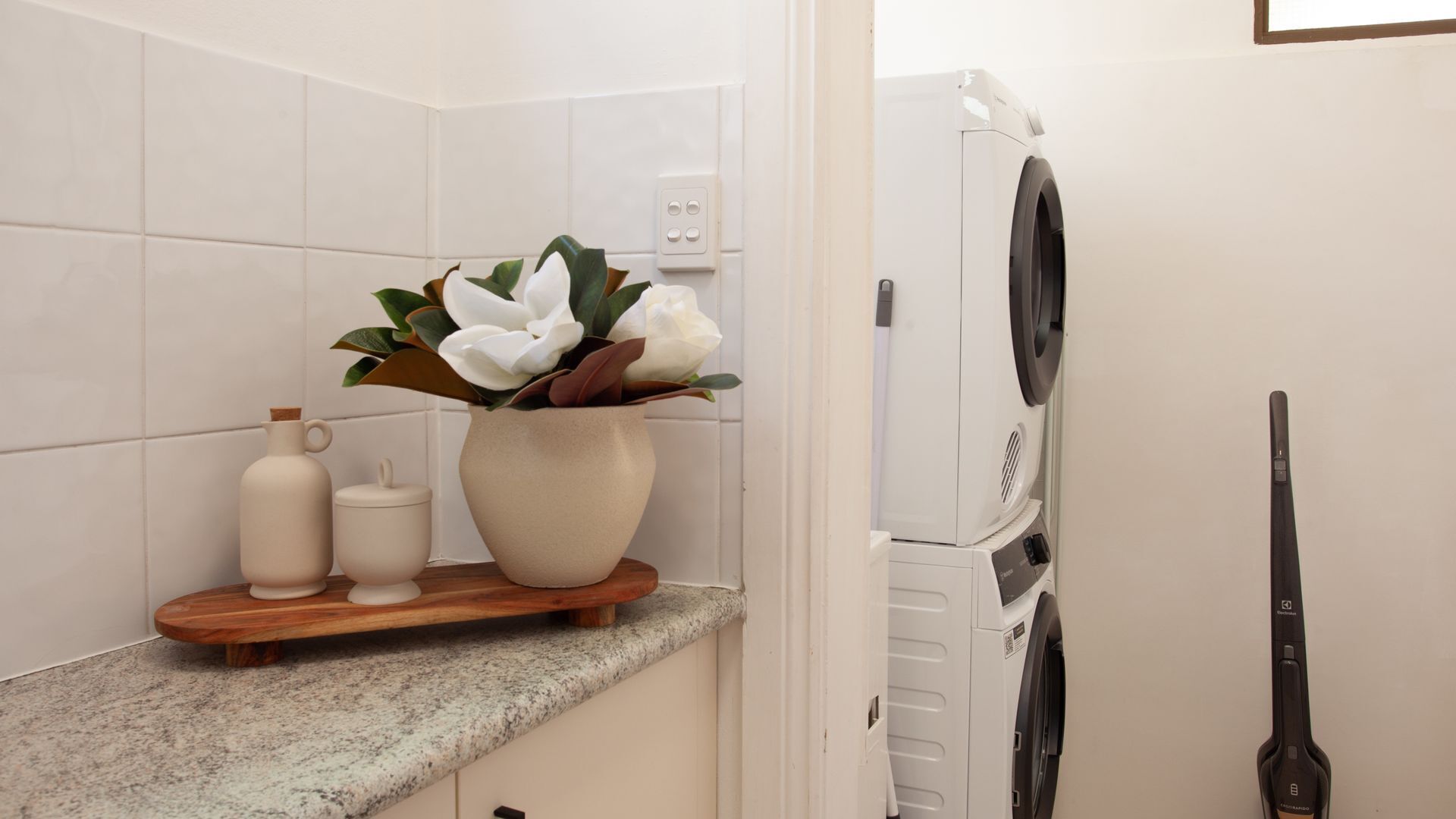 A bathroom with a white tiled wall, a wooden shelf with a vase of flowers, a white washing machine, and a hairdryer.