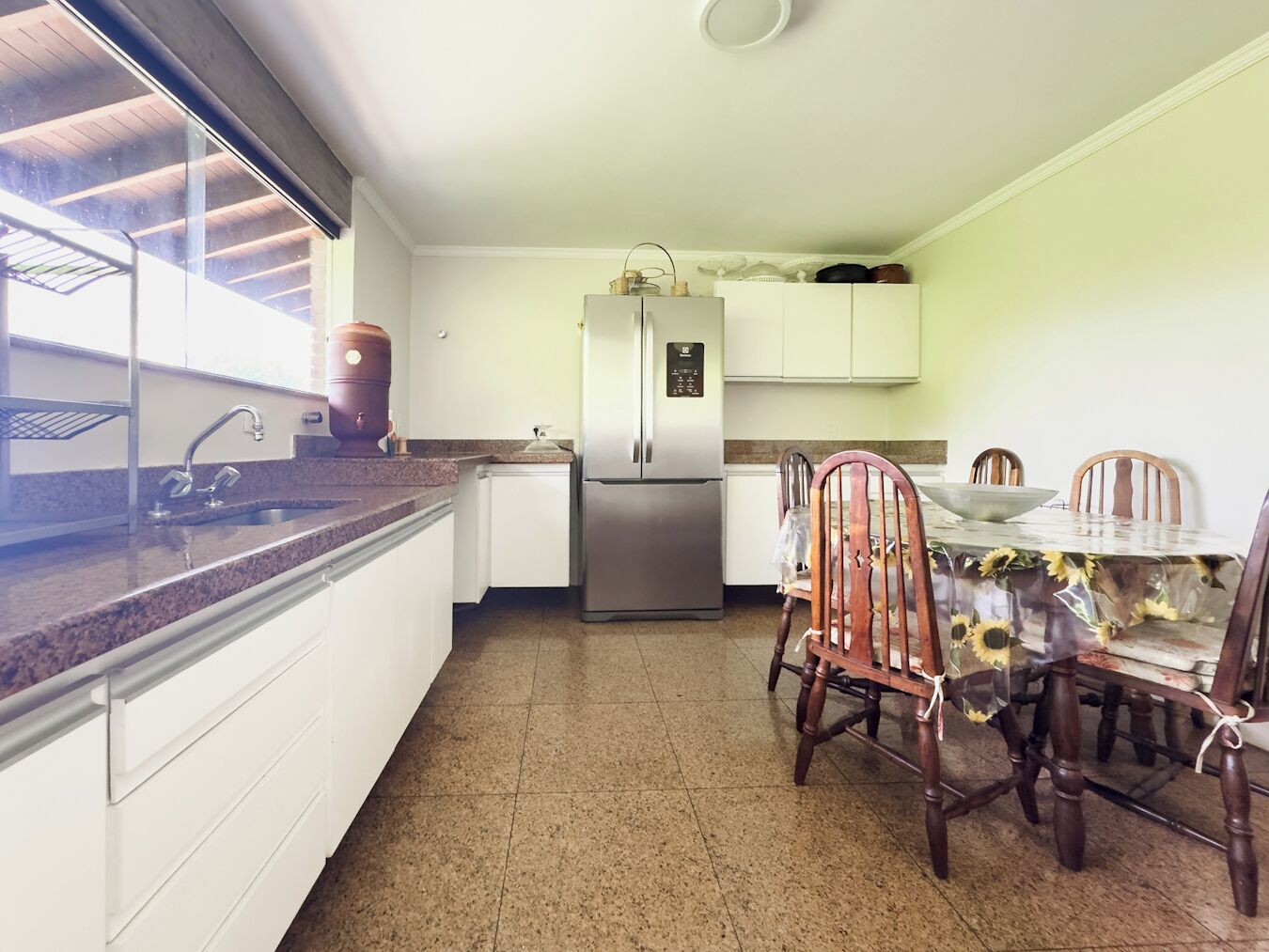 Uma cozinha compacta com armários brancos, uma geladeira de aço inoxidável, bancada de granito e uma área de jantar com cadeiras e mesa de madeira.
