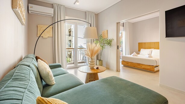 A modern hotel room with a green sofa, a round wooden table with a lamp, a bed with white linens, and a large window. - Acropolis Path - Elegant Apartment near Plaka (Athina)