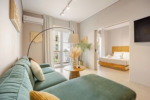 A modern hotel room with a green sofa, a round wooden table with a lamp, a bed with white linens, and a large window. - Acropolis Path - Elegant Apartment near Plaka (Athina)