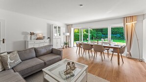 A modern living room with a sofa, coffee table, dining area with chairs, and a large window with a view of greenery.