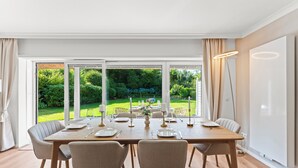 A dining area with a large table set for six, surrounded by chairs, with a view of a garden through large windows.