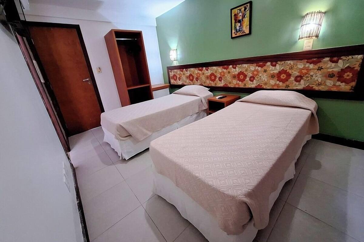 Ein Hotelzimmer mit zwei Betten, einem Holzschrank, einem Kopfteil mit Blumenmuster und einem kleinen Tisch mit Lampe.