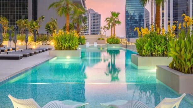 A rooftop pool with clear blue water, surrounded by palm trees and plants, and modern buildings in the background. - Modern Studio Near Dubai Mall, Walk to Dubai Canal (Dubai)