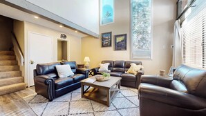 A living room with a staircase, a coffee table with decorative items, and a large window with a view of trees. - Modern Cabin for 8, Near Slopes + Canyon Lodge! (Mammoth Lakes)