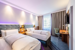 Ett hotellrum med två sängar, ett skrivbord, en stol, en TV och ett fönster med gardiner.