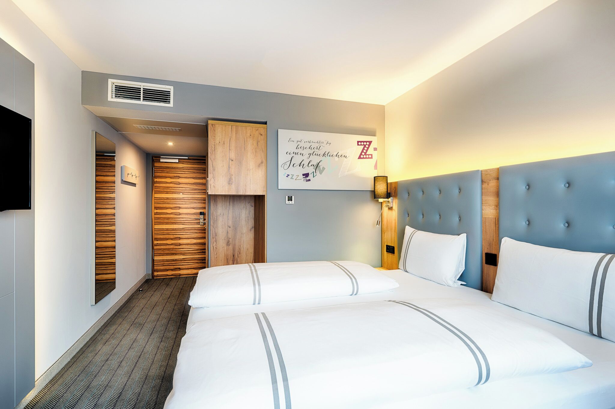 A hotel room with a large bed, a wooden wardrobe, a wall-mounted artwork, and a ceiling vent.