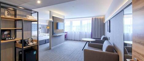 Et moderne hotelværelse med en seng, et siddeområde med sofa og et lille bord, minibar samt et fjernsyn.