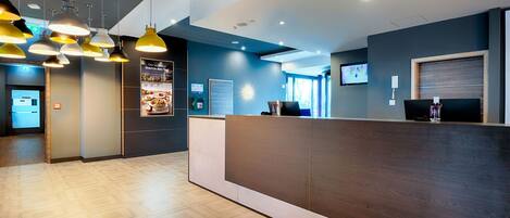 A hotel reception area with a wooden reception desk, multiple pendant lights, a digital screen, and a poster on the wall.
