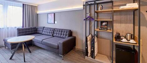 Una habitación de hotel moderna con un sofá seccional gris, una mesa redonda, un armario empotrado con estantes y un refrigerador negro pequeño.
