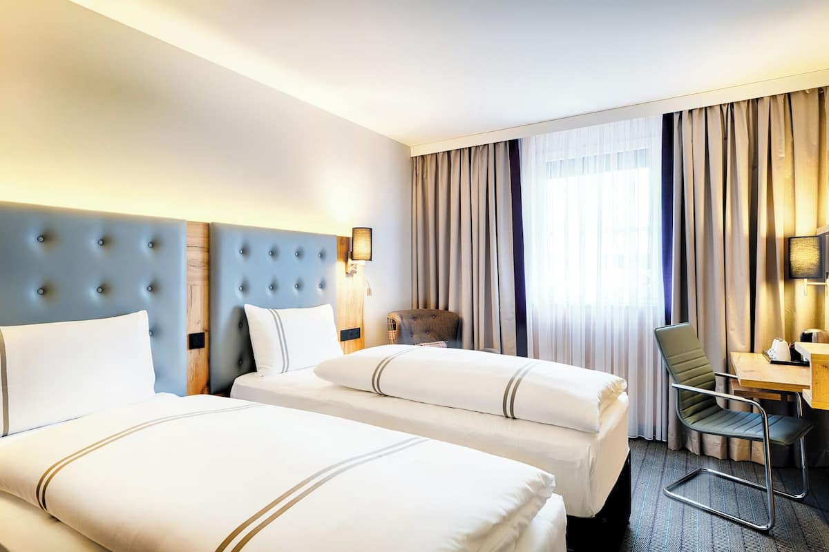 A hotel room with two beds, a desk, a chair, and a large window with curtains.