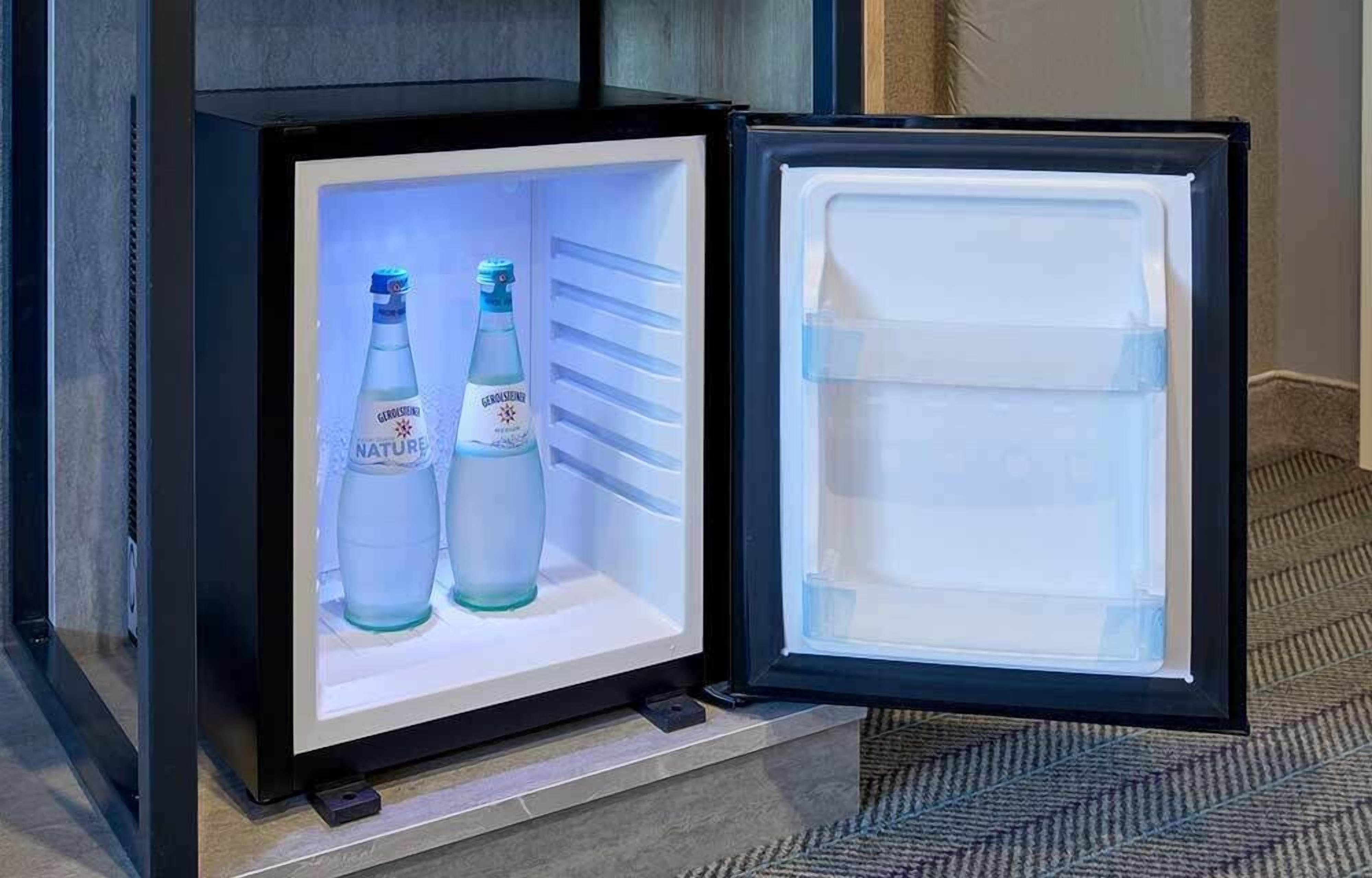 A mini-fridge with a built-in display shelf containing two bottles of Gerolsteiner Nature water and a glass jar.
