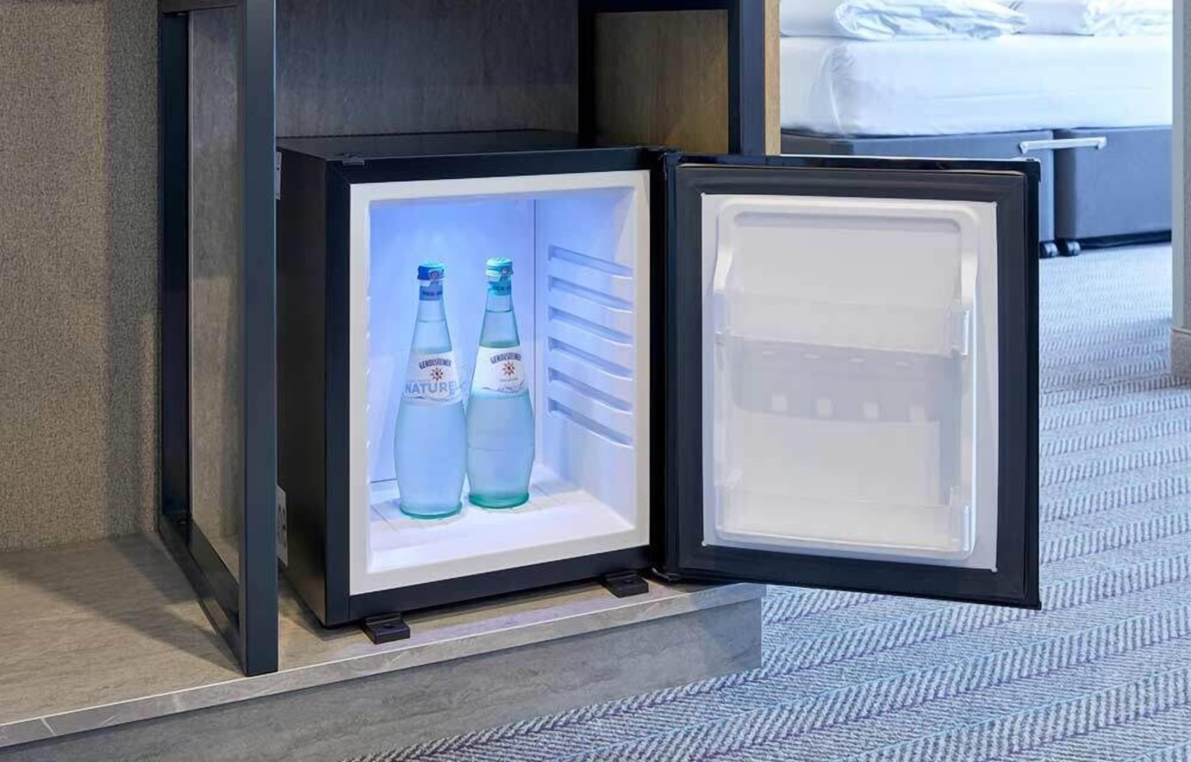 A compact refrigerator with two bottles of water inside, placed on a tiled floor near a bed.